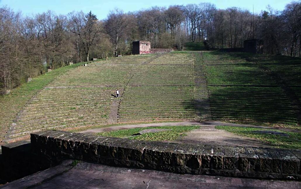 A view of the huge seating area of the Heidelberg Thingstatte, looking out from the stage. A person walking up the steps among the seats shows the enormous scale. Trees behind.