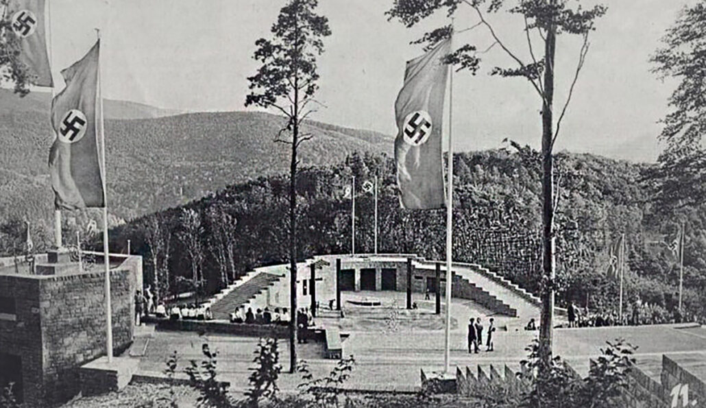 A black-and-white vintage photo, ca. 1935, looking down onto the Thingstatte stage, with Nazi flags flying from several poles. A few people are walking around the stage area.