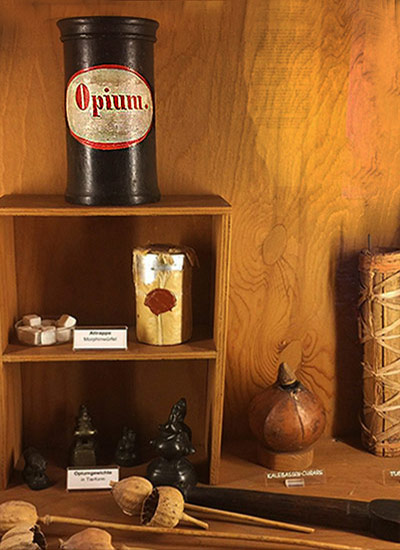 Medicaments in a showcase including a black jar with a white and red label "Opium." Also dried poppy seed pods on the table.