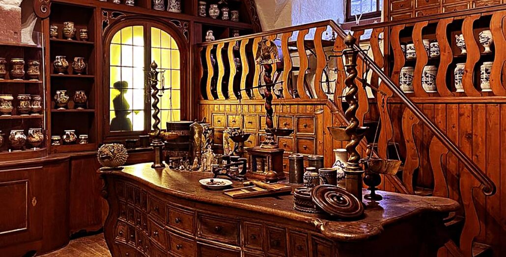 The Bamberg Court Pharmacy at the German Apothecary Museum. We see the wood worktable strewn with various tools and materials, plus a large pharmacist's scale. Behind the table is a sort of mezzanine with steps up to a desk where the apothecary could do paperwork. On the left wall is a center-opening multi-paned window with the silhouette of a woman customer behind it, waiting to speak to the pharmacist. The woodwork has a warm golden glow.